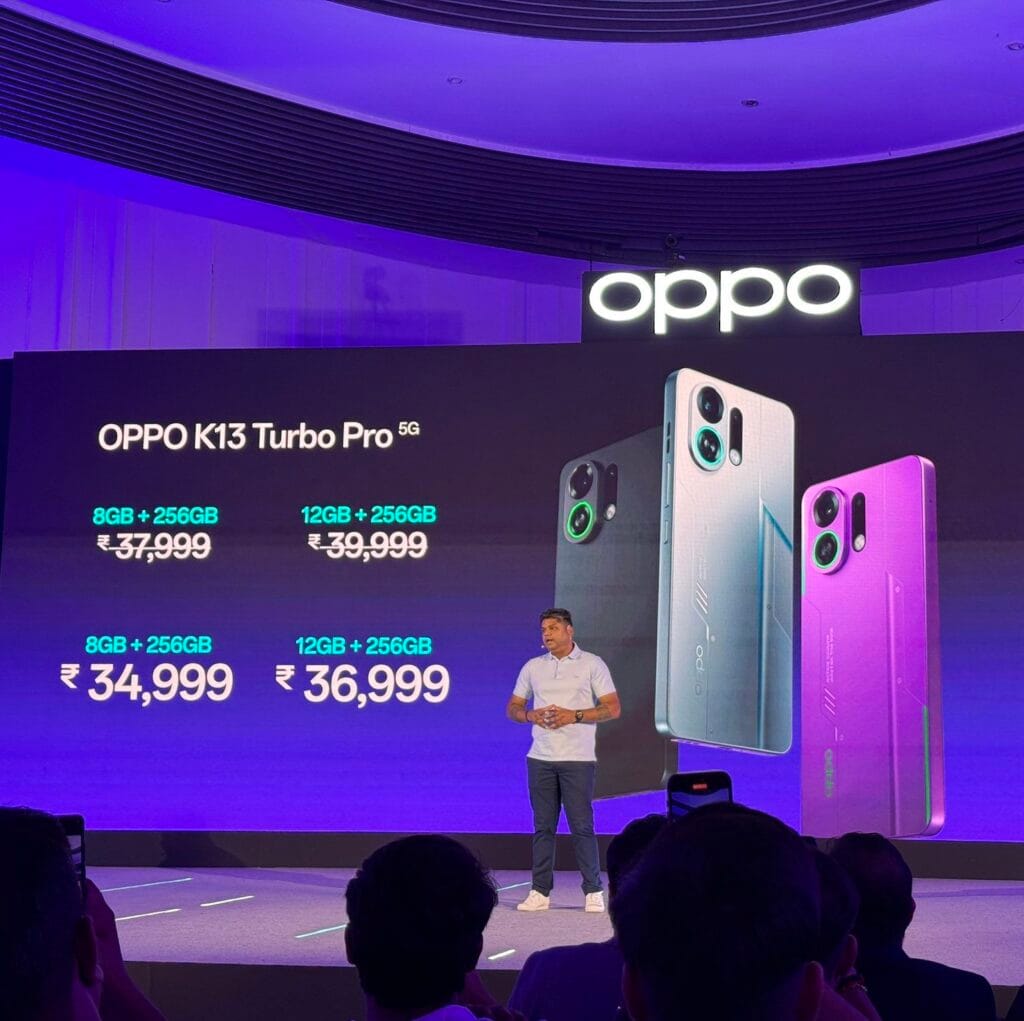 Oppo K13 Turbo, K13 Turbo Pro Officially Launched in India 2 A presentation stage with a large screen displaying the Oppo K13 Turbo Pro 5G prices: 8GB+256GB for ₹37,999 and 12GB+256GB for ₹39,999. Below, it shows "8GB+256GB ₹34,999" and "12GB+256GB ₹36,999". A presenter stands on stage. Three phone models in dark gray, white, and purple are displayed next to the prices. The Oppo logo is visible at the top. Audience members are in the foreground.