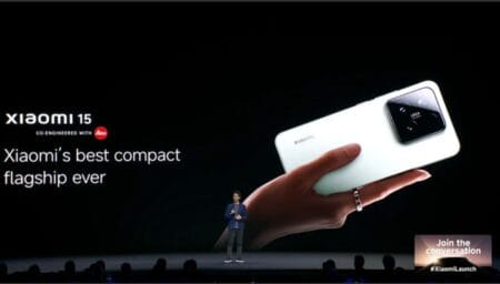 A live event showcasing the Xiaomi 15 5G. A presenter stands center stage as a large screen displays a close-up of the phone held in a hand, along with the tagline "Xiaomi's best compact flagship ever." The phone is light green and co-engineered with Leica. The hashtag #XiaomiLaunch is visible.