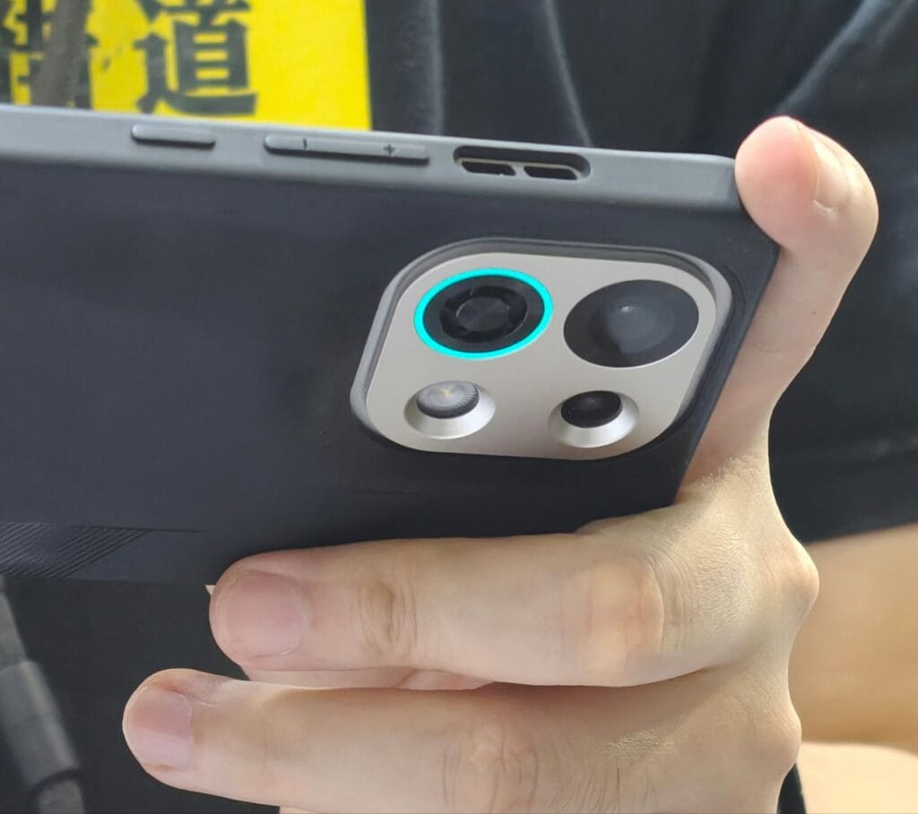 Oppo K13 Turbo | Built-in Active Cooling Fan 2 Close-up of a person holding the Oppo K13 Turbo smartphone, showcasing its rear camera module with a distinctive teal accent ring around the main lens. The phone is in a dark-colored case.