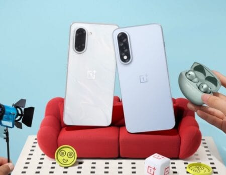Two OnePlus Nord 5 smartphones, one white and one light blue, are shown leaning against each other on a small red couch. A pair of OnePlus earbuds in a case rests nearby. The scene is playfully styled with miniature props.