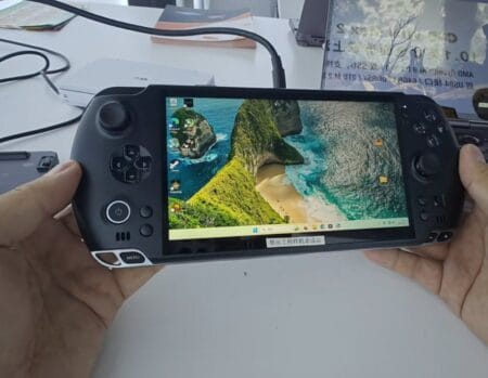 Close-up of a GPD Win 5 handheld console held in two hands. The screen shows a desktop environment with a tropical beach wallpaper. The device has gaming controls on either side of the screen.