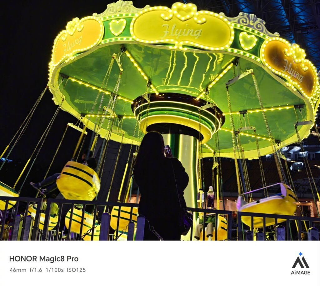 A nighttime photograph captured with HONOR Magic8 Pro (46mm, f/1.6, 1/100s, ISO125) showcases a brightly illuminated swing ride. The ride features a green canopy adorned with yellow lights, heart motifs, and the word "Flying" on its upper edge. Yellow swing seats hang below, while a person in dark clothing stands in the foreground taking a photo. The image includes HONOR Magic8 Pro branding and the "AIMAGE" logo at the bottom.