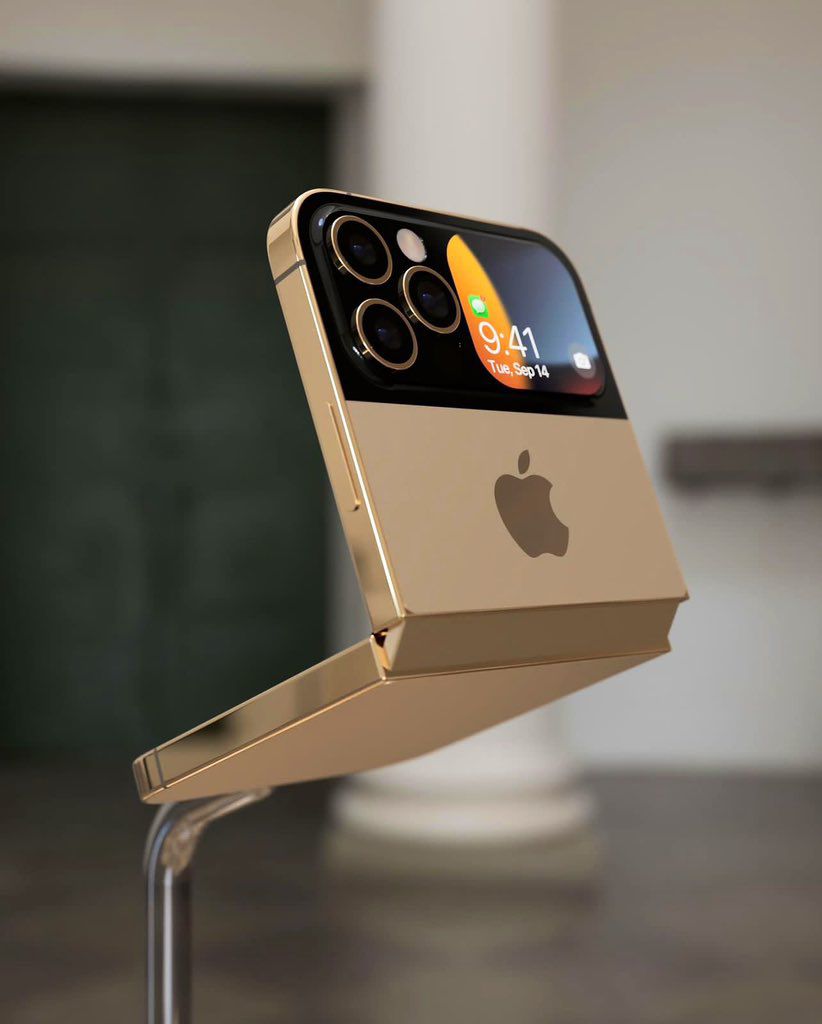 A gold-colored foldable iPhone with four cameras and a small screen on the back, showing the time and date. The phone is resting on a surface and the background is blurred.