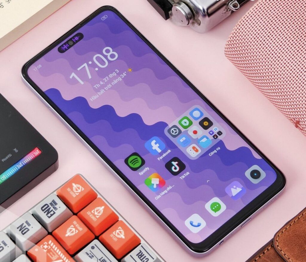 A Realme C65 smartphone with a purple wavy-pattern wallpaper and various app icons on the home screen, placed on a desk amidst colorful accessories and gadgets.