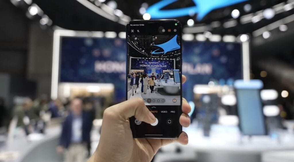 Honor Magic 7 Pro Arrives in PH on April 10, 2025 2 Close-up of a hand holding a smartphone, likely an Honor phone, in camera mode. The phone's screen shows a photo of a trade show booth with the text 'Honor Alpha Plan'.