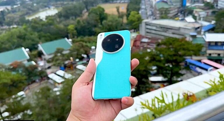 Close-up view of a hand holding the Tecno Camon 40 Premier 5G smartphone. The phone is teal with a unique circular camera module on the back. The background is blurred, showing a cityscape.