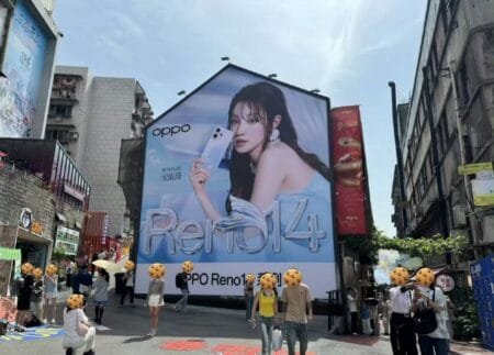 Oppo Reno 14 (PKZ110) Spotted on Geekbench (China) with MediaTek Dimensity 8450 A vibrant street scene with a massive billboard dominating the view. The billboard showcases the Oppo Reno 14 series phone with an image of actress Song Yuqi. Several pedestrians are visible, with some partially obscured, walking past the advertisement in a bustling city street.