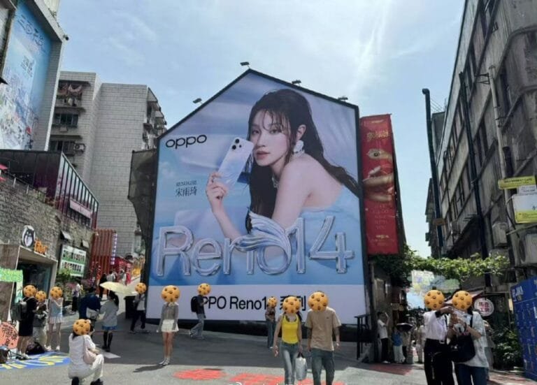 A vibrant street scene with a massive billboard dominating the view. The billboard showcases the Oppo Reno 14 series phone with an image of actress Song Yuqi. Several pedestrians are visible, with some partially obscured, walking past the advertisement in a bustling city street.