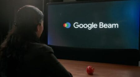 A woman sits facing a large screen displaying the Google Beam logo. A red apple rests on the table in front of her.