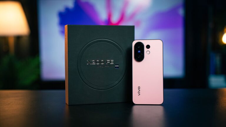A pink Vivo X200 FE smartphone rests on a dark surface next to its dark gray box.  The box features the phone's name in metallic lettering. A blurred background shows a computer screen with a colorful image.