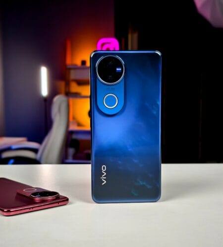 The rear camera module of a blue Vivo V50 smartphone is prominently displayed. A partially visible red phone is in the foreground. The background is out of focus, suggesting an indoor setting.