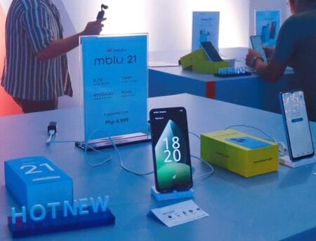 The Meizu mBlu 21 smartphone is showcased at a product launch event. The phone sits on a display stand alongside its packaging and promotional materials, highlighting key features such as its large screen and suggested price of PHP 4,999. Other Meizu products are also visible in the background.