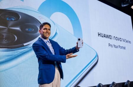 HUAWEI Nova 14 Launched in the Philippines, Priced at ₱22,999 A man in a blue suit presents the Huawei nova 14 at its launch event. The phone is silver with a large circular camera module.