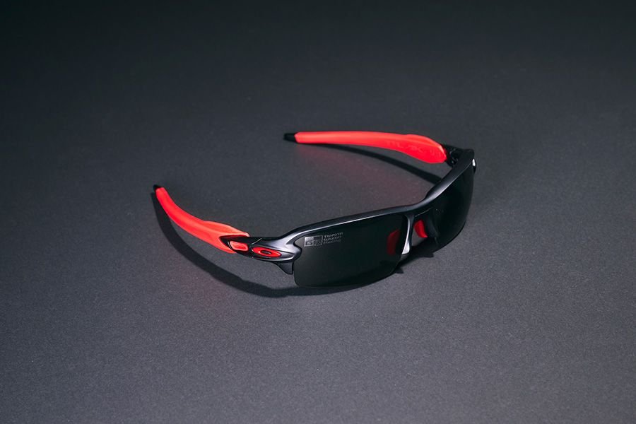 Pair of collaborative sunglasses from Toyota GAZOO Racing (TGR) and Oakley, launching at Tokyo Auto Salon 2026. The sports-style frames feature a matte black front and bright red temple arms, with dark tinted lenses. Displayed against a plain dark gray background.