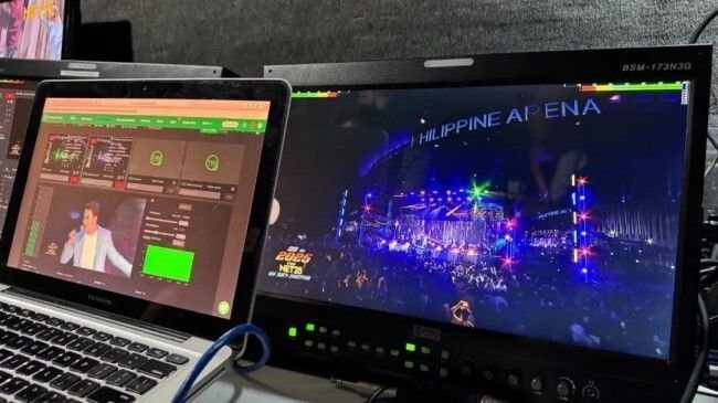 Two screens display content from the Philippine Arena event. The laptop on the left shows a broadcast control interface with video feeds and green status indicators. The larger monitor on the right (labeled 'BSM-173N3G') shows a live view of the arena’s stage with bright blue and colorful lighting, and 'PHILIPPINE ARENA' text at the top.