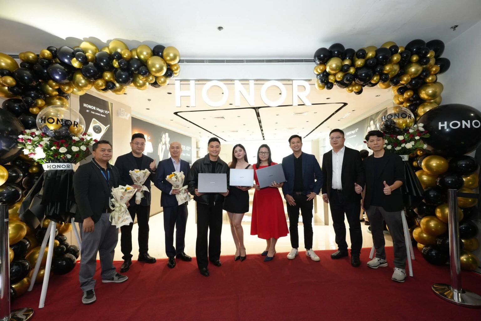 A group of people stand in front of an HONOR store, decorated with black and gold balloons, holding laptops and bouquets, celebrating the opening of HONOR Philippines' 200th store.