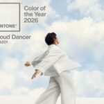 Pantone Color of the Year 2026 announcement featuring 'Cloud Dancer' (11-4201). A person in flowing white clothing stands against a backdrop of soft, cloud-filled sky, evoking tranquility.