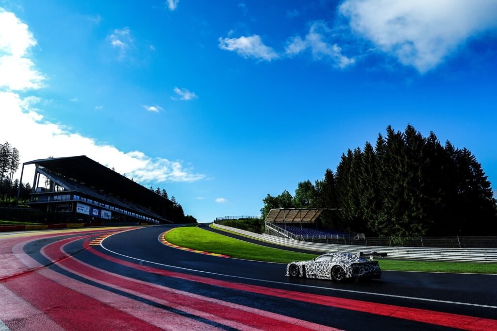 Toyota Gazoo Racing’s New GR GT3 Turns Heads as It Gears Up for Racetracks and Roads 1 A camouflaged Toyota GR GT3 prototype speeds through a scenic racetrack under a bright blue sky, framed by grandstands and lush greenery.