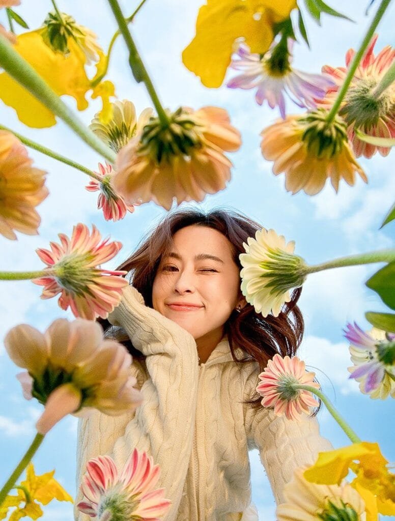 A low-angle, close-up shot of a woman with long brown hair, wearing a cream-colored cable-knit sweater. She is surrounded by a profusion of colorful flowers, which appear slightly blurred and out-of-focus, creating a dreamy, whimsical effect. The sky is visible in the background. This is a camera sample of asus zenfone 12 ultra.