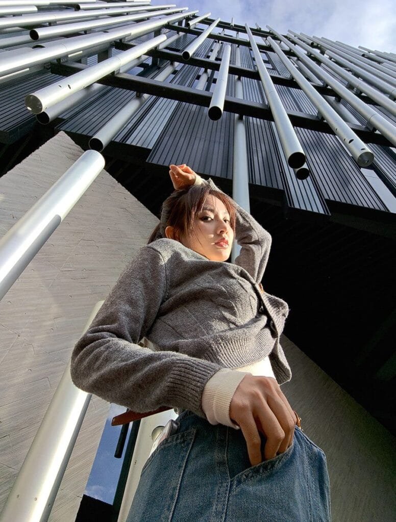 A low-angle, full shot of a young woman with dark hair, wearing a grey cardigan and blue jeans, standing in front of a building with a striking facade of vertical metal pipes. The woman is posing with one hand on her head and the other in her jeans pocket. The image is taken from a perspective looking upward towards the building and the sky is visible. This is a camera sample of asus zenfone 12 ultra