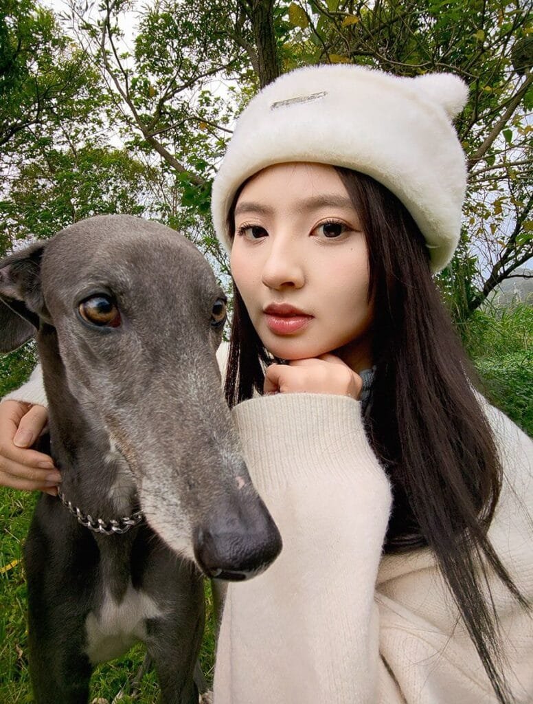 A close-up selfie shows a young woman with long dark hair wearing a fluffy white hat. She is gently holding a greyhound dog, which is positioned next to her face. The background is slightly blurred, showing green foliage and trees. This is a camera sample of asus zenfone 12 ultra