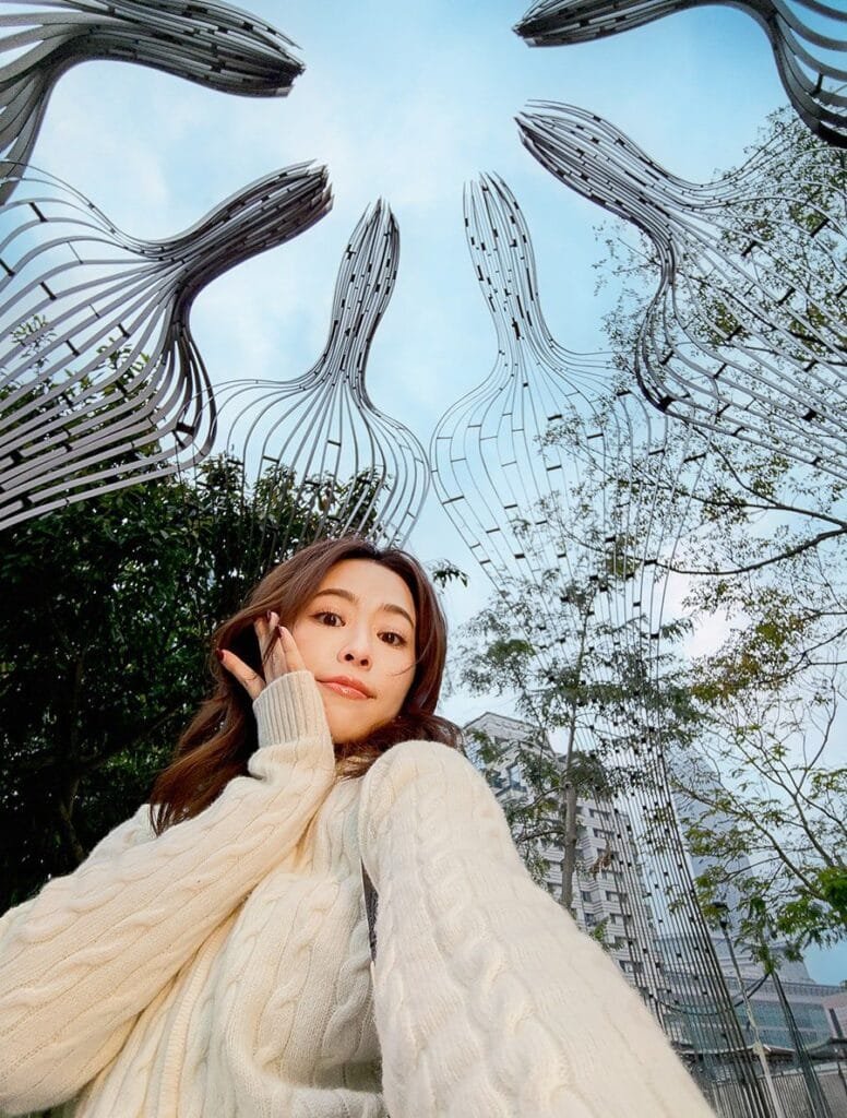 Low-angle selfie of a woman with long brown hair, wearing a cream-colored cable-knit sweater.  She's posing with one hand near her face. In the background are several tall, modern, metallic sculptures that arch overhead against a partly cloudy sky. This is a camera sample of asus zenfone 12 ultra