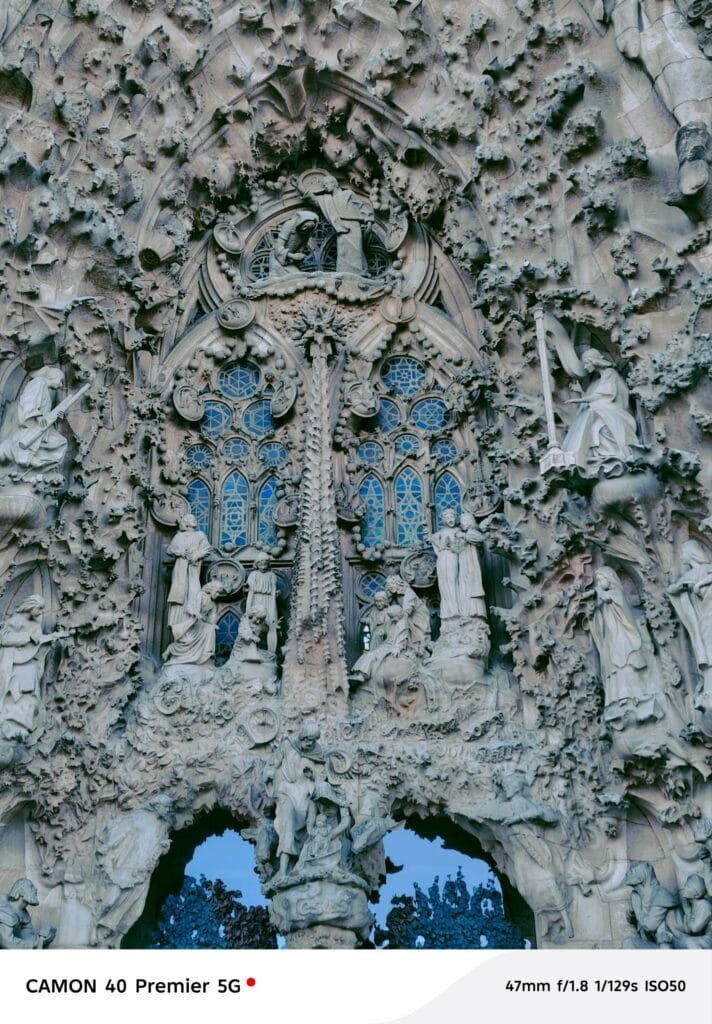 A close-up photograph showcasing the exquisite detail of a section of the Sagrada Familia's facade in Barcelona. The image highlights the numerous sculpted figures and the stained-glass window within the elaborate stonework. Photographed with a Tecno Camon 40 Premier 5G.