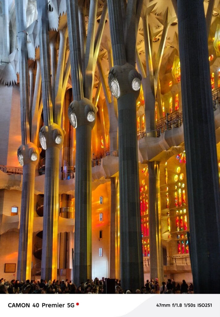 A photograph of the interior of the Sagrada Familia basilica in Barcelona, Spain. The image focuses on the forest of slender, uniquely shaped columns supporting the soaring ceiling, and the vibrant stained-glass windows illuminating the space. Photographed with a Tecno Camon 40 Premier 5G.
