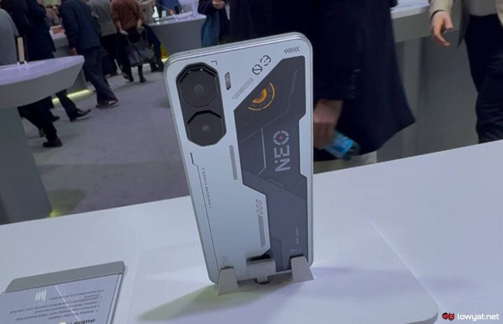 Close-up view of the back of a Nubia Neo 3 5G smartphone. The phone is predominantly white and silver with black accents, featuring a distinctive design with angular lines and the word "NEO" prominently displayed. The camera module is a noticeable feature, with two large lenses. The phone is displayed upright on a small stand against a blurred background showing people in what appears to be a trade show or product launch setting.