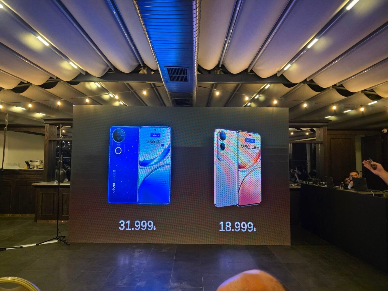 Vivo V50 Lite 5G Coming Soon to PH: Massive 6500mAh Bluevolt Battery w/ 90W Fast Charge A large screen at a product launch event displays the Vivo V50 and Vivo V50 Lite smartphones. The Vivo V50 is shown in blue, and the Vivo V50 Lite in a rose gold color. Prices are displayed beneath each phone in Turkish Lira (₺31,999 and ₺18,999 respectively).