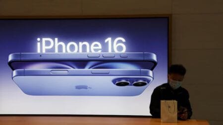 A large display shows an image of the iPhone 16 in a store in Indonesia. A person in the foreground holds an Apple shopping bag, suggesting the recent lifting of a sales ban on the phone.