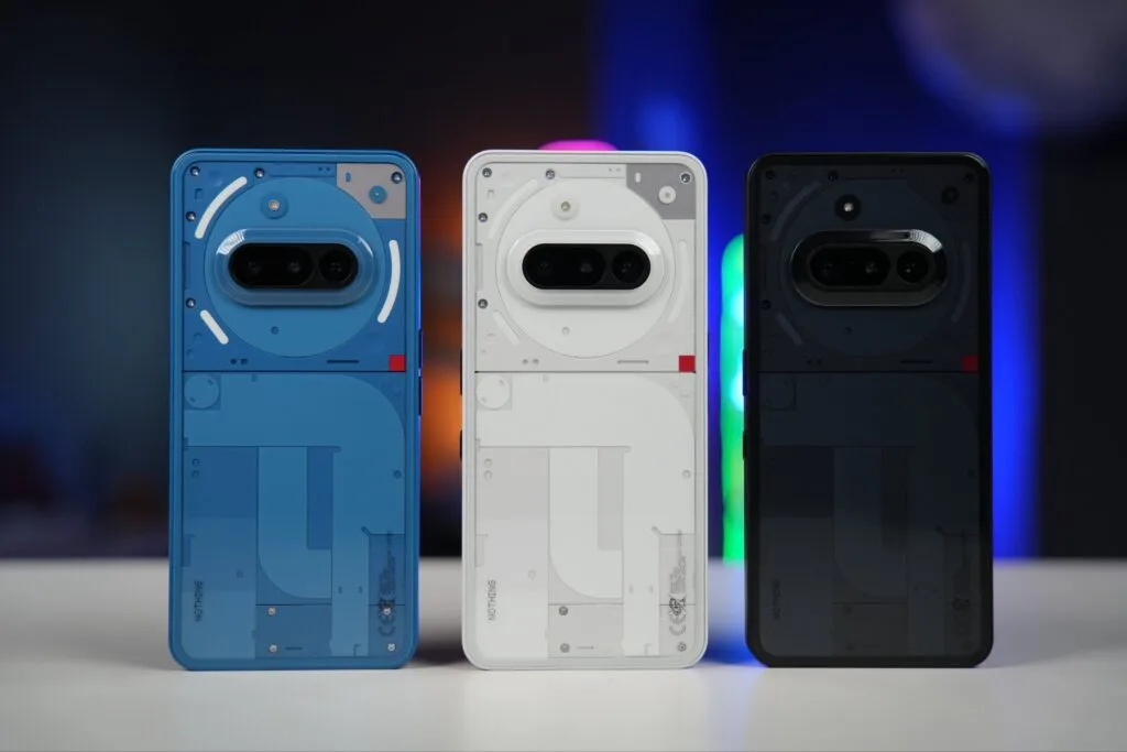 Three Nothing Phone (3a) smartphones are displayed, showcasing their transparent backs revealing internal components. The phones are positioned side-by-side on a white surface, with a blurred background of colorful lights. The phones are in blue, white, and black.