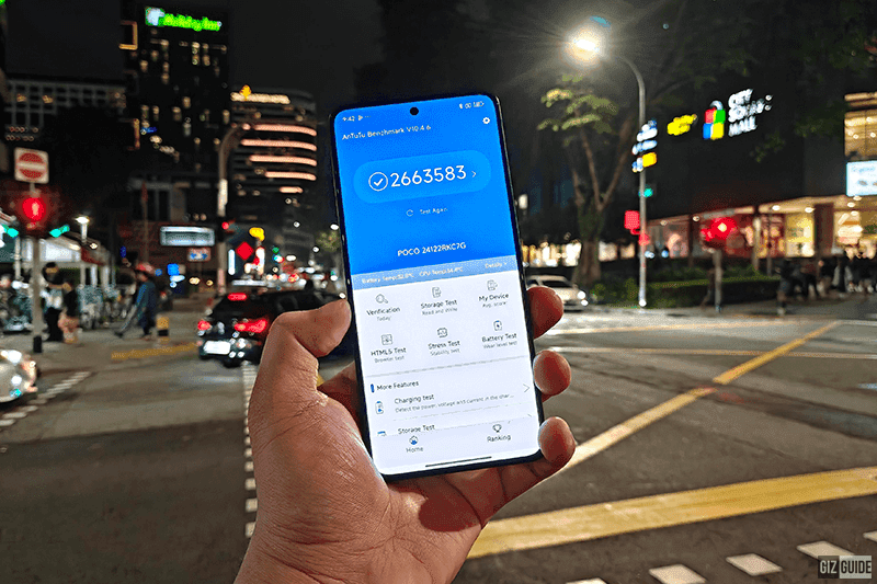 ‎A hand holds a smartphone displaying the Antutu Benchmark app. The screen shows a score of 2663583 and the device name "POCO 241228KC7G". The background is a blurry night scene of a city street with traffic lights and a building in the distance. The text "GizGuide" appears at the bottom right corner of the image.