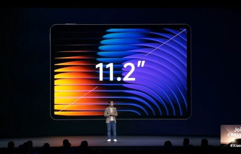 A photograph of a product launch event of Xiaomi Pad 7 series. A large screen dominates the image, displaying the number "11.2" in a large, sans-serif font. The background behind the number is a vibrant abstract graphic featuring curved lines in shades of orange, yellow, blue, and purple. A man in a light brown blazer and jeans is standing on a stage in front of the screen, seemingly presenting the product. A small portion of an audience is visible in the lower part of the image, and a small graphic in the lower right corner suggests a call to action or social media hashtag. The overall scene suggests a technology product reveal.