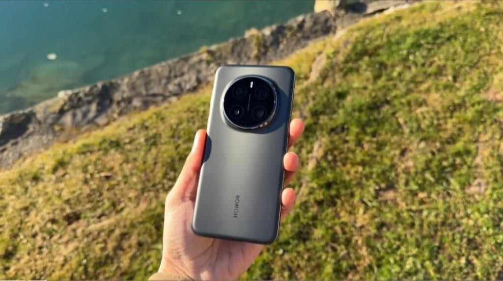 ‎A hand holds up an Honor Magic 7 Pro smartphone, showcasing its back and circular camera design. The phone is gray and the background is blurred, showing a grassy area near water.