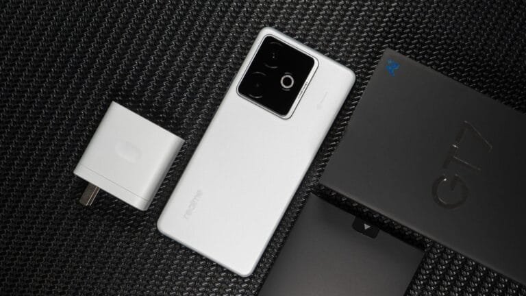 A white Realme GT 7 smartphone lies on a black textured surface next to its charger and retail box.