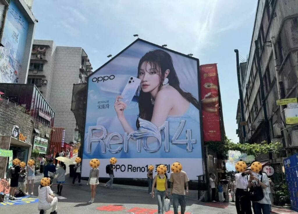 A vibrant street scene with a massive billboard dominating the view. The billboard showcases the Oppo Reno 14 series phone with an image of actress Song Yuqi. Several pedestrians are visible, with some partially obscured, walking past the advertisement in a bustling city street.