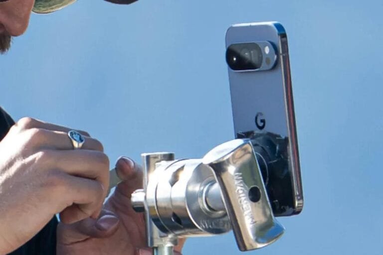 A person's hand adjusts a silver Google Pixel 10 series smartphone attached to a professional-looking tripod. The phone's rear camera system is visible. The background is a blurred-out light blue sky.
