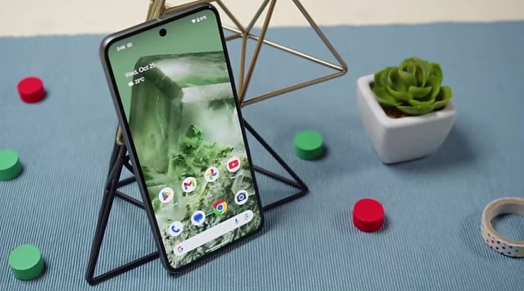 A Google Pixel phone, possibly a Pixel 8, is displayed on a stand. The phone's screen shows the date "Wed, May 7, 2025," The screen also displays several Google apps icons. The phone rests on a blue surface with small decorative objects nearby.