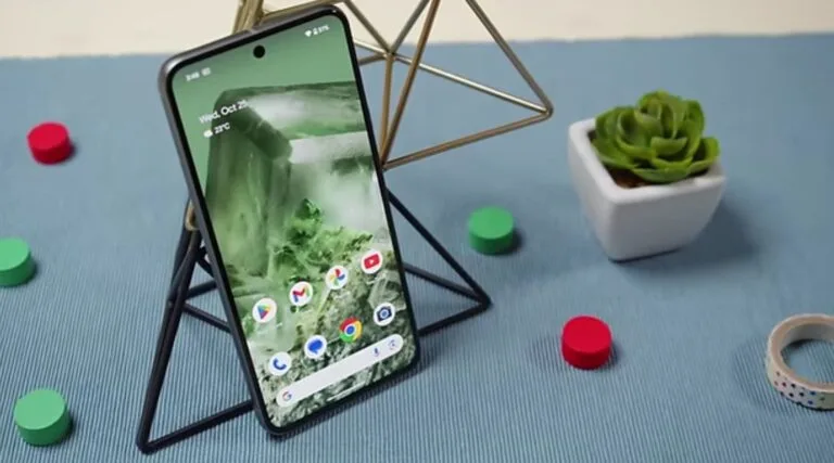 List of Google Pixel Updates: May 2025 Rollout Begins—Mic Recording & Bluetooth Pairing A Google Pixel phone, possibly a Pixel 8, is displayed on a stand. The phone's screen shows the date "Wed, May 7, 2025," The screen also displays several Google apps icons. The phone rests on a blue surface with small decorative objects nearby.