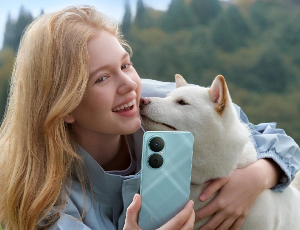 Close-up of a young woman with long blonde hair smiling and cuddling a white Shiba Inu dog. She's holding up a light teal Huawei Nova Y73 phone to take a selfie. The background is a softly blurred green landscape.