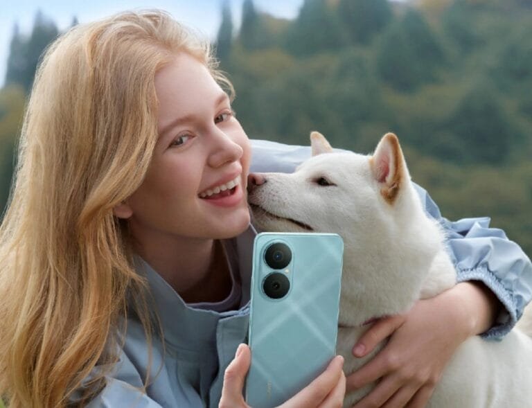 Close-up of a young woman with long blonde hair smiling and cuddling a white Shiba Inu dog. She's holding up a light teal Huawei Nova Y73 phone to take a selfie. The background is a softly blurred green landscape.