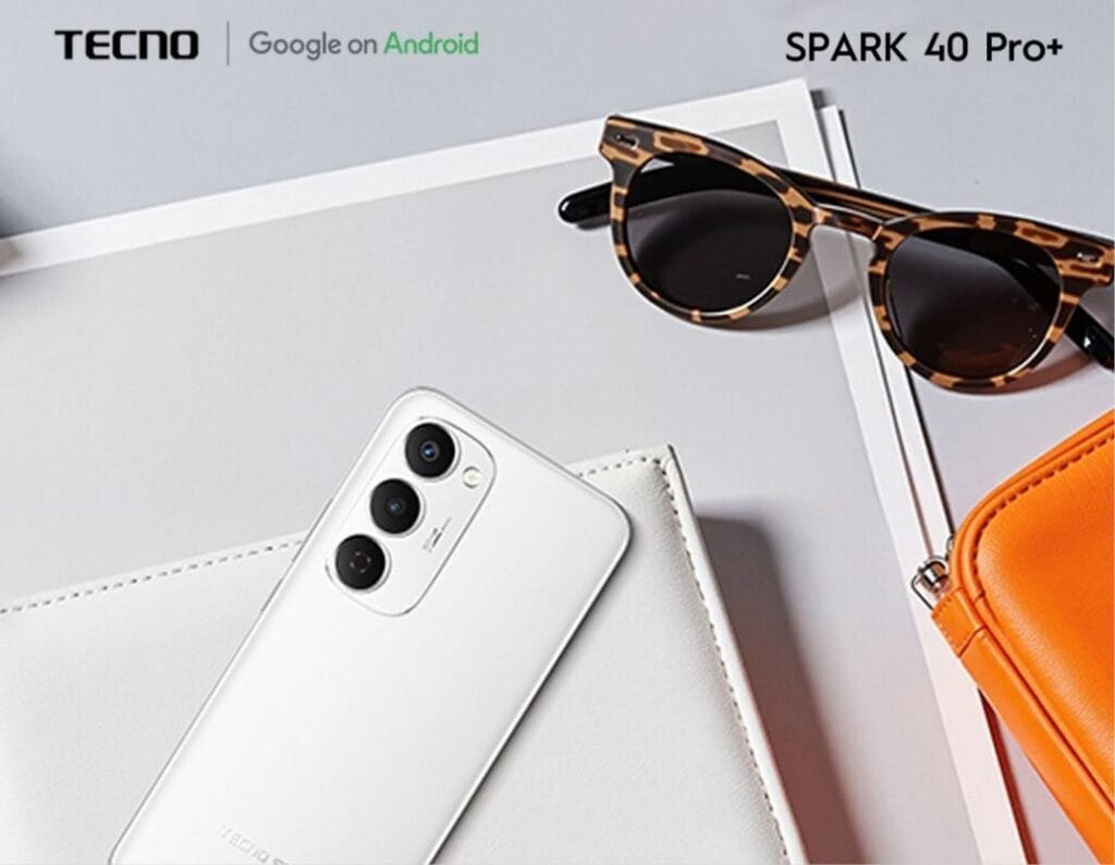 A white Tecno Spark 40 Pro+ smartphone rests on a light gray surface next to tortoiseshell sunglasses and an orange pouch. The phone's camera is clearly visible.
