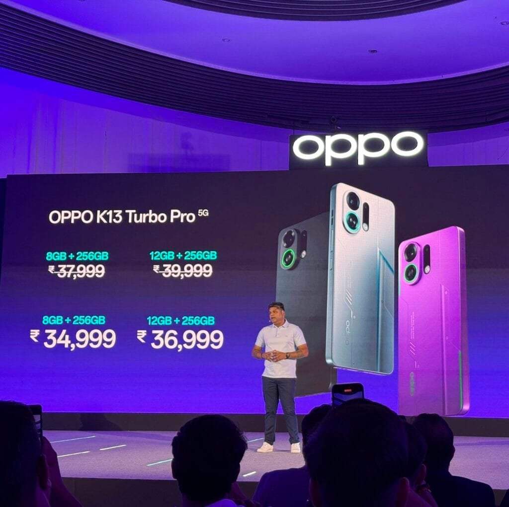 A presentation stage with a large screen displaying the Oppo K13 Turbo Pro 5G prices: 8GB+256GB for ₹37,999 and 12GB+256GB for ₹39,999. Below, it shows "8GB+256GB ₹34,999" and "12GB+256GB ₹36,999". A presenter stands on stage. Three phone models in dark gray, white, and purple are displayed next to the prices. The Oppo logo is visible at the top. Audience members are in the foreground.
