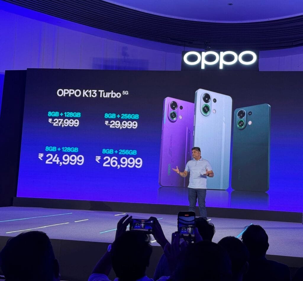 A presentation stage with a large screen displaying the Oppo K13 Turbo 5G prices: 8GB+128GB for ₹27,999 and 8GB+256GB for ₹29,999. Below, it shows "8GB+128GB ₹24,999" and "8GB+256GB ₹26,999". A presenter stands on stage, gesturing towards the screen. Three phone models in purple, light blue, and dark green are displayed next to the prices. The Oppo logo is visible at the top. Audience members are taking photos with their phones.