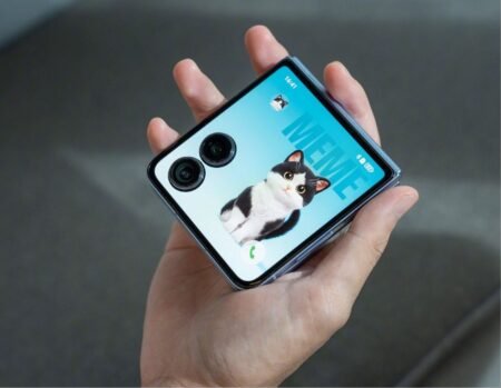 A hand holds the Honor Magic V Flip 2, showcasing its dual rear cameras and a screen displaying a cat meme wallpaper.