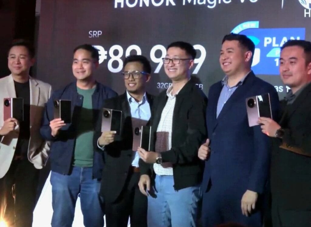 Six men in business attire pose with the Honor Magic V5 at its Philippine launch event. The backdrop displays the phone's SRP and partnership with Globe Telecom.