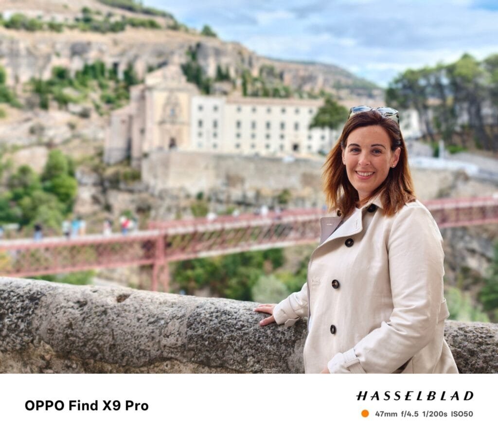 Oppo Find X9 Pro portrait: Woman in a trench coat smiling with a bridge and buildings in the background.