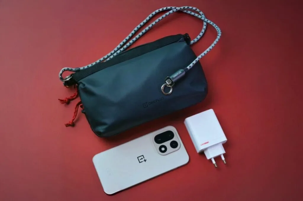 OnePlus 15 sand dune edition on a red surface, showcasing its design alongside a charger and a stylish OnePlus crossbody bag.