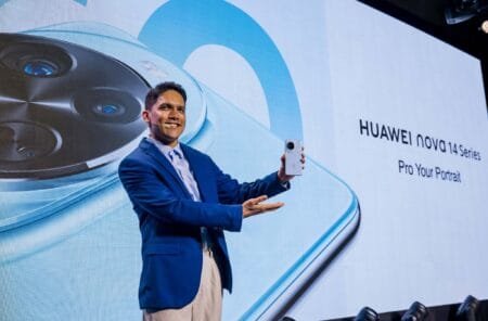 A man in a blue suit presents the Huawei nova 14 at its launch event. The phone is silver with a large circular camera module.